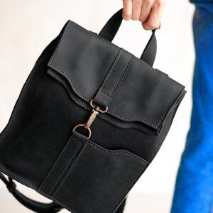 Pode incluir: Uma mochila de couro preto fosco com al&ccedil;a superior, aba dobr&aacute;vel com detalhes de costura e um fecho central de mosquet&atilde;o de metal em tom de lat&atilde;o antigo. A bolsa inclui um bolso frontal e al&ccedil;as de ombro ajust&aacute;veis.
