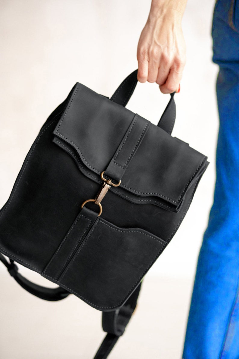 Pode incluir: Uma mochila de couro preto fosco com al&ccedil;a superior, aba dobr&aacute;vel com detalhes de costura e um fecho central de mosquet&atilde;o de metal em tom de lat&atilde;o antigo. A bolsa inclui um bolso frontal e al&ccedil;as de ombro ajust&aacute;veis.