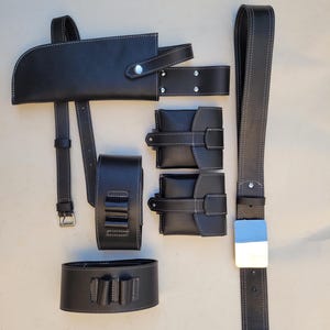 Pu&ograve; includere: Un set di accessori in pelle nera per una cintura, tra cui una fondina, due tasche e un braccialetto. Gli accessori hanno cuciture bianche e dettagli argentati.