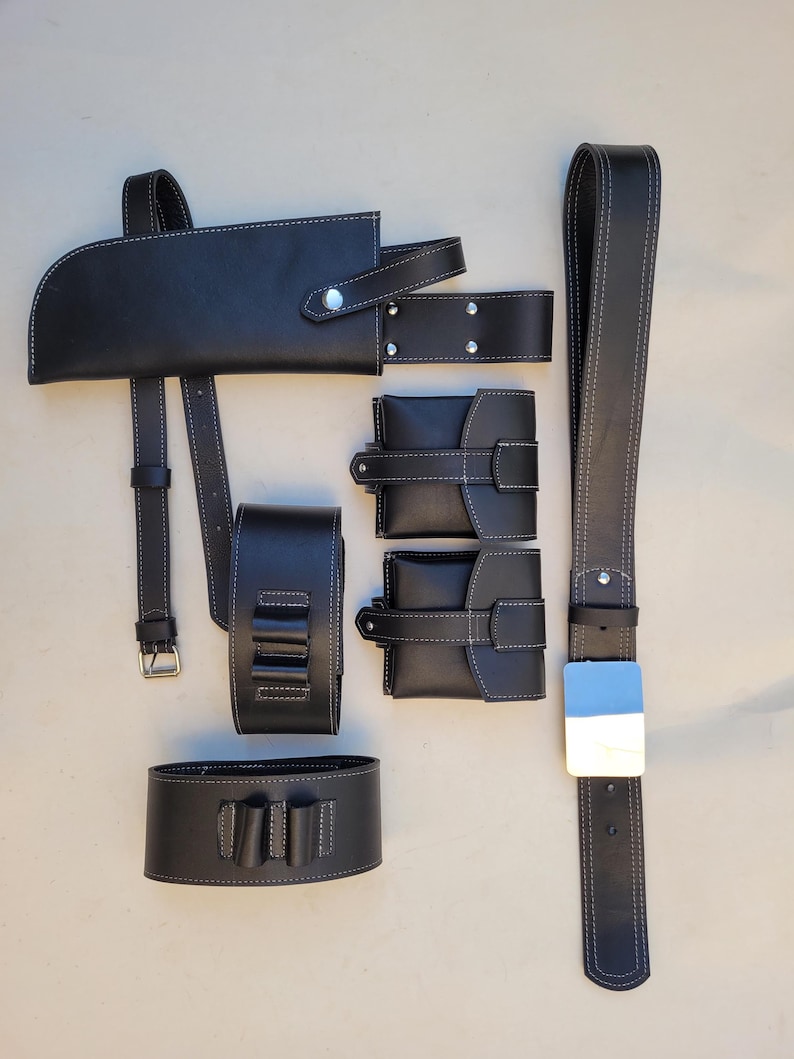 Pu&ograve; includere: Un set di accessori in pelle nera per una cintura, tra cui una fondina, due tasche e un braccialetto. Gli accessori hanno cuciture bianche e dettagli argentati.