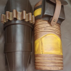 Może przedstawiać: Dwa brązowe sk&oacute;rzane ochraniacze na ramiona ze szlufkami i klamrami. Jeden ochraniacz ma wz&oacute;r w ż&oacute;łto-brązowe pasy i małą brązową kieszeń z paskiem.