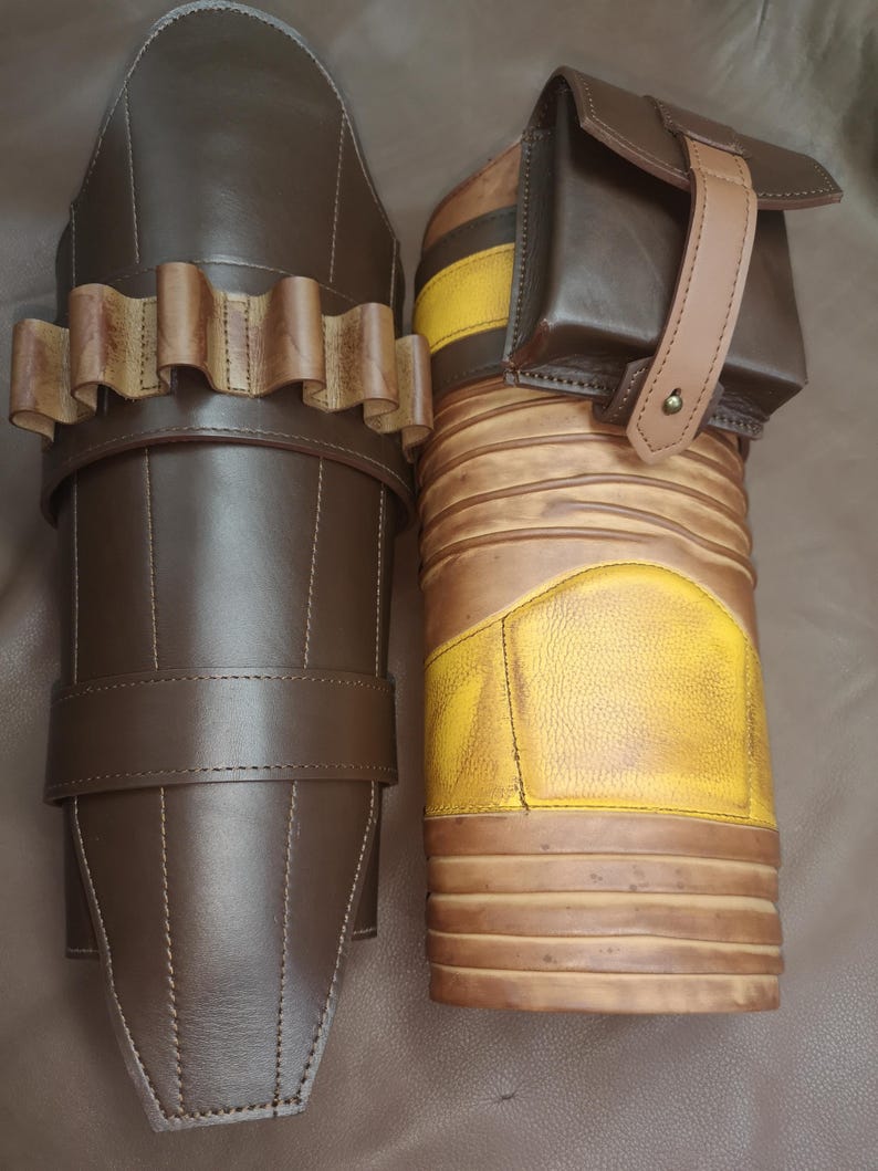 Może przedstawiać: Dwa brązowe sk&oacute;rzane ochraniacze na ramiona ze szlufkami i klamrami. Jeden ochraniacz ma wz&oacute;r w ż&oacute;łto-brązowe pasy i małą brązową kieszeń z paskiem.