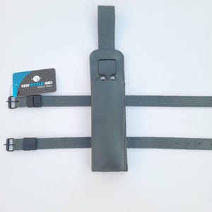 Peut inclure: Un étui de téléphone en cuir gris avec une sangle et une boucle. L'étui a une boucle en haut pour l'accrocher. Le texte "TEN STYLE IND" est visible sur une étiquette blanche.