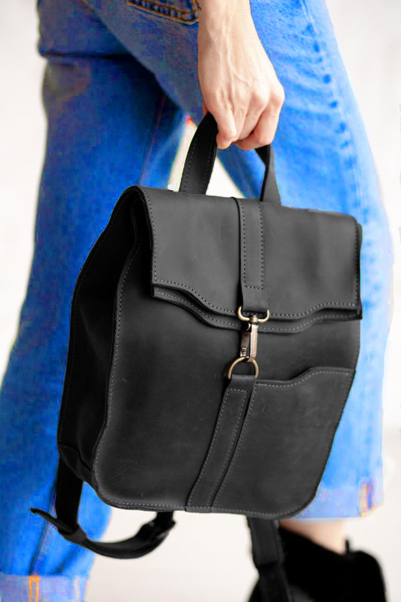 Pode incluir: Uma mochila de couro preto fosco com al&ccedil;a superior, aba com uma tira vertical central e fecho de mosquet&atilde;o met&aacute;lico. A bolsa inclui um bolso frontal e al&ccedil;as de ombro ajust&aacute;veis com costuras vis&iacute;veis em toda a sua estrutura.