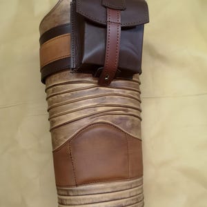 Może przedstawiać: Brązowa sk&oacute;rzana kabura na nogę z mniejszą, brązową sk&oacute;rzaną kieszenią. Kabura ma ozdobny wz&oacute;r i brązowy sk&oacute;rzany pasek.