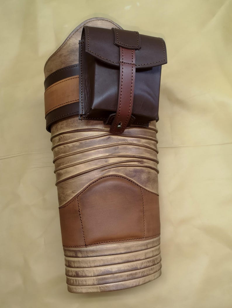 Może przedstawiać: Brązowa sk&oacute;rzana kabura na nogę z mniejszą, brązową sk&oacute;rzaną kieszenią. Kabura ma ozdobny wz&oacute;r i brązowy sk&oacute;rzany pasek.