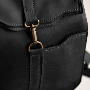 Pode incluir: Uma mochila de couro preto com acabamento fosco, costuras tonais vis&iacute;veis e um fecho de mosquet&atilde;o de metal cor de lat&atilde;o preso a uma al&ccedil;a de couro vertical. A bolsa inclui um bolso lateral com abertura curva e bordas refor&ccedil;adas.
