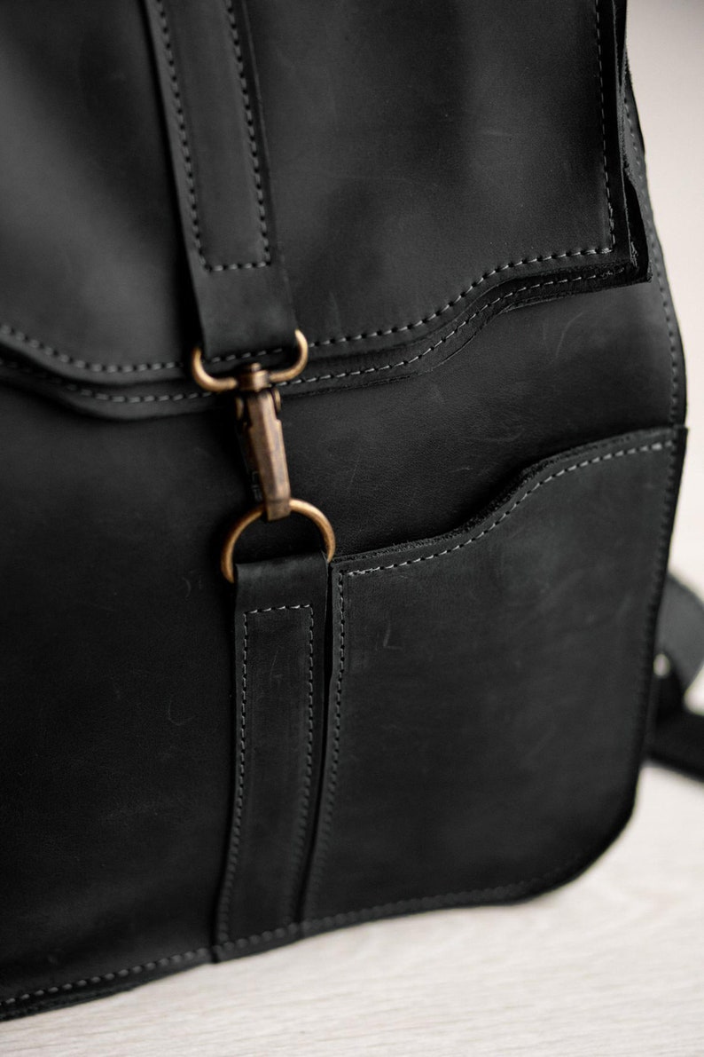 Pode incluir: Uma mochila de couro preto com acabamento fosco, costuras tonais vis&iacute;veis e um fecho de mosquet&atilde;o de metal cor de lat&atilde;o preso a uma al&ccedil;a de couro vertical. A bolsa inclui um bolso lateral com abertura curva e bordas refor&ccedil;adas.