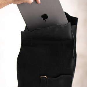 Pode incluir: Uma mochila de couro preto fosco com um bolso frontal vertical e um fecho em forma de anel em D na cor lat&atilde;o. Um tablet cinza-escuro com o log&oacute;tipo da Apple vis&iacute;vel est&aacute; a ser inserido no compartimento principal da mala estruturada.