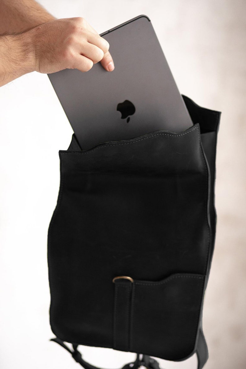 Pode incluir: Uma mochila de couro preto fosco com um bolso frontal vertical e um fecho em forma de anel em D na cor lat&atilde;o. Um tablet cinza-escuro com o log&oacute;tipo da Apple vis&iacute;vel est&aacute; a ser inserido no compartimento principal da mala estruturada.