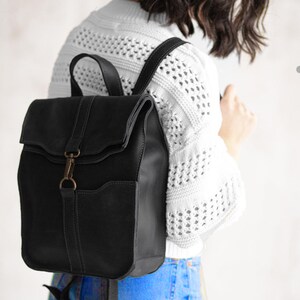 Pode incluir: Uma mochila de couro preta com fecho de aba dobr&aacute;vel fixado com um gancho de metal e fecho de anel em tom de bronze. A bolsa inclui uma al&ccedil;a superior, al&ccedil;as de ombro ajust&aacute;veis e um bolso frontal com detalhes de costura vis&iacute;veis.