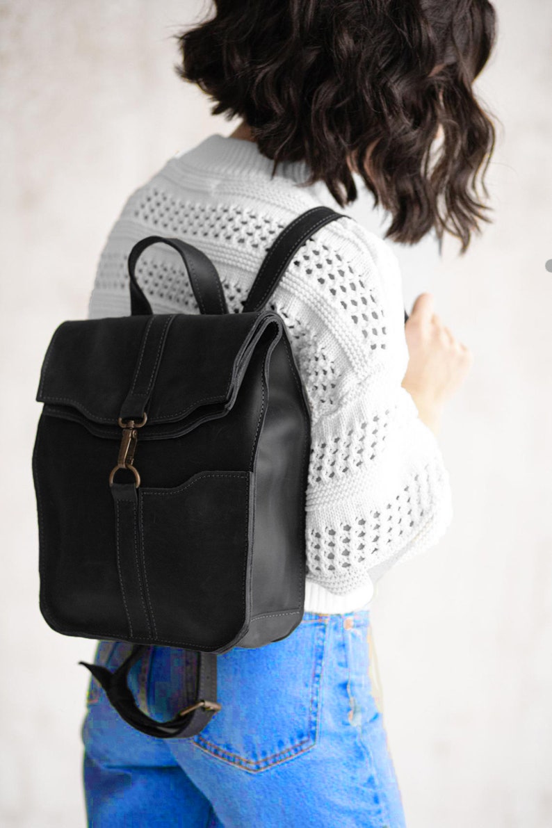 Pode incluir: Uma mochila de couro preta com fecho de aba dobr&aacute;vel fixado com um gancho de metal e fecho de anel em tom de bronze. A bolsa inclui uma al&ccedil;a superior, al&ccedil;as de ombro ajust&aacute;veis e um bolso frontal com detalhes de costura vis&iacute;veis.