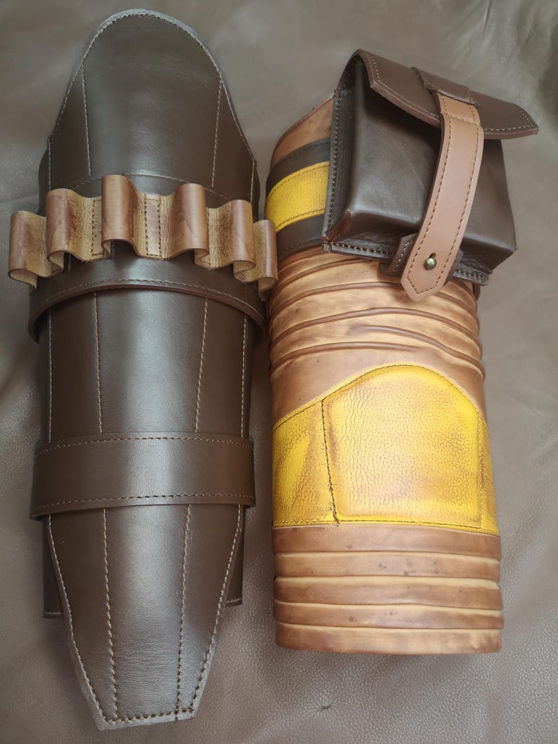 Może przedstawiać: Para brązowych sk&oacute;rzanych ochraniaczy na nogi z ż&oacute;łto-brązowym paskowanym wzorem. Jeden ochraniacz ma małą brązową sk&oacute;rzaną kieszeń z paskiem.