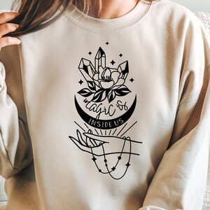 Puede incluir: Una sudadera beige con un diseño gráfico negro de una mano sosteniendo una luna creciente con cristales y una flor. El texto "Magic Is Inside Us" está impreso en la sudadera.