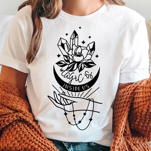 Puede incluir: Camiseta blanca con un gráfico en blanco y negro de una mano sosteniendo una luna creciente con cristales y una flor. El texto "Magic is Inside Us" está impreso debajo del gráfico.