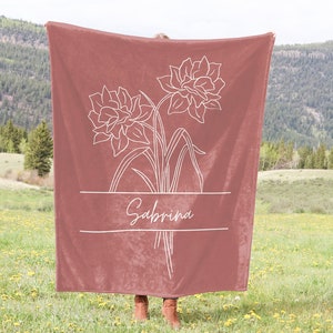 May include: A dusty rose blanket featuring a white floral design and the name "Sabrina" in a script font. The blanket is held up in a field with a mountain backdrop. The floral design includes two large flowers and stems.