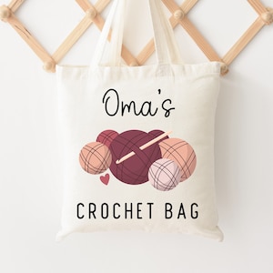 May include: Cream-colored tote bag with the text "Oma's CROCHET BAG" in black script. The bag features a graphic of yarn balls in shades of burgundy and peach, with a crochet hook. The bag is hanging on a wooden wall-mounted rack.
