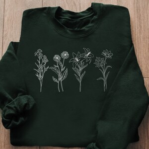 Pode incluir: Uma camisa de moletom verde escura com um contorno branco de v&aacute;rias flores na frente.