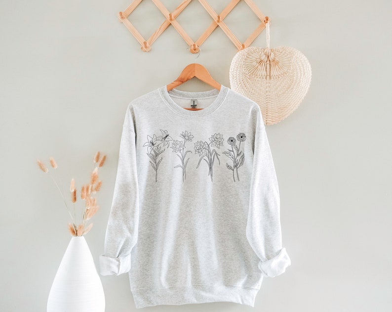 Pode incluir: Uma camisa de moletom cinza com um desenho de linha preta de uma variedade de flores na frente.