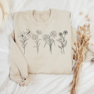 Pode incluir: Uma camisa de moletom bege com um desenho de linha preta de cinco flores diferentes na frente.