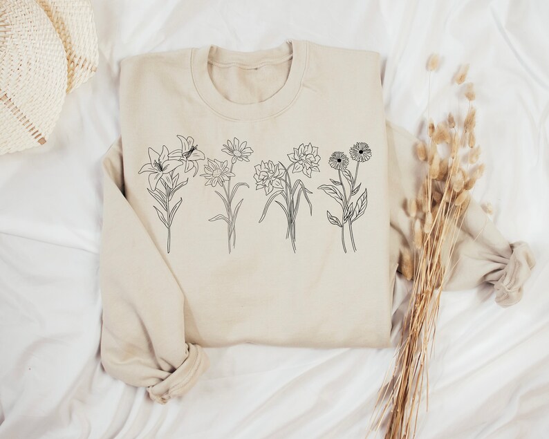 Pode incluir: Uma camisa de moletom bege com um desenho de linha preta de cinco flores diferentes na frente.