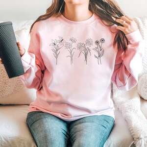 Pode incluir: Uma camisa de moletom rosa com um desenho de flores em preto na frente.