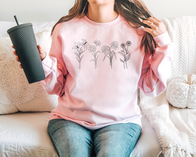 Pode incluir: Uma camisa de moletom rosa com um desenho de flores em preto na frente.