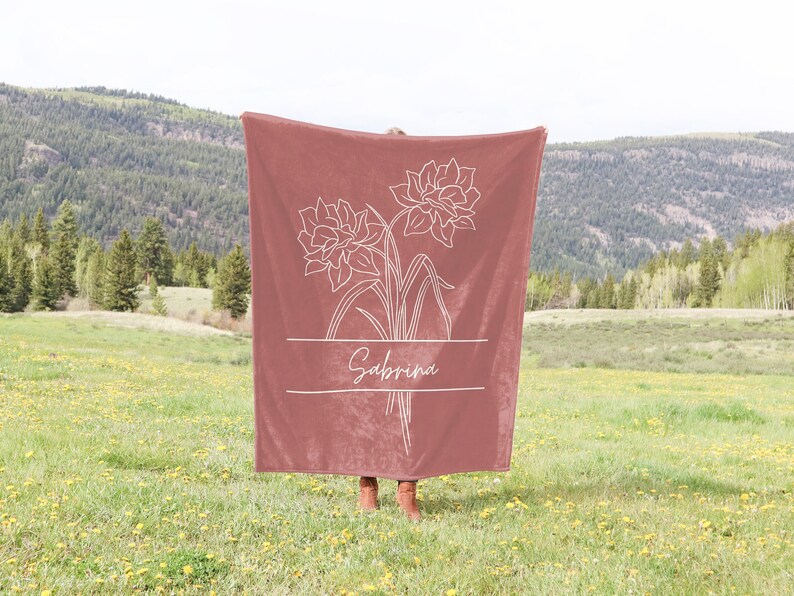 May include: A dusty rose blanket featuring a white floral design and the name "Sabrina" in a script font. The blanket is held up in a field with a mountain backdrop. The floral design includes two large flowers and stems.