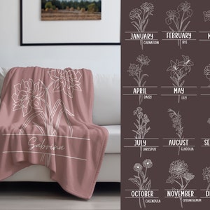 May include: A dusty rose throw blanket with a white floral design and the name "Sabrina" in a white rectangular box. The blanket is draped over a white sofa. A chart of birth month flowers is on the right.