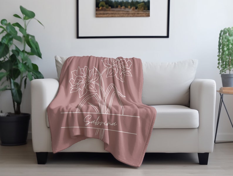 May include: A dusty rose throw blanket draped over a white sofa. The blanket features a white floral design and the name "Sabrina" in a white font. A framed picture hangs on the wall behind the sofa, and a potted plant is to the left.