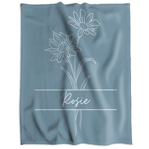 May include: A blue-grey towel featuring a white floral line drawing with two daisies and the name "Rosie" in cursive script. The design is centered on the towel, with the name placed below the flowers within a rectangular frame.