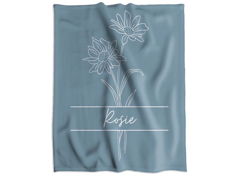 May include: A blue-grey towel featuring a white floral line drawing with two daisies and the name "Rosie" in cursive script. The design is centered on the towel, with the name placed below the flowers within a rectangular frame.