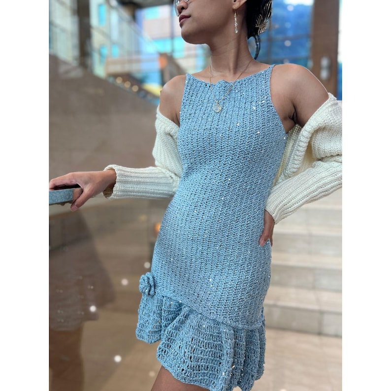 Puede incluir: Un vestido de crochet azul claro con un dobladillo con volantes y una chaqueta blanca. El vestido es sin mangas y tiene una silueta ajustada.