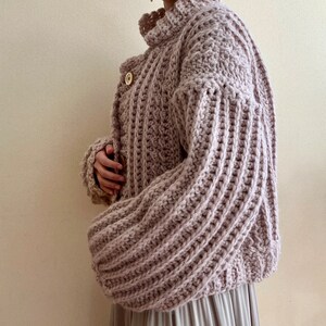 May include: A light pink crocheted cardigan with a high neck and long sleeves. The cardigan has a button closure and a textured stitch pattern.