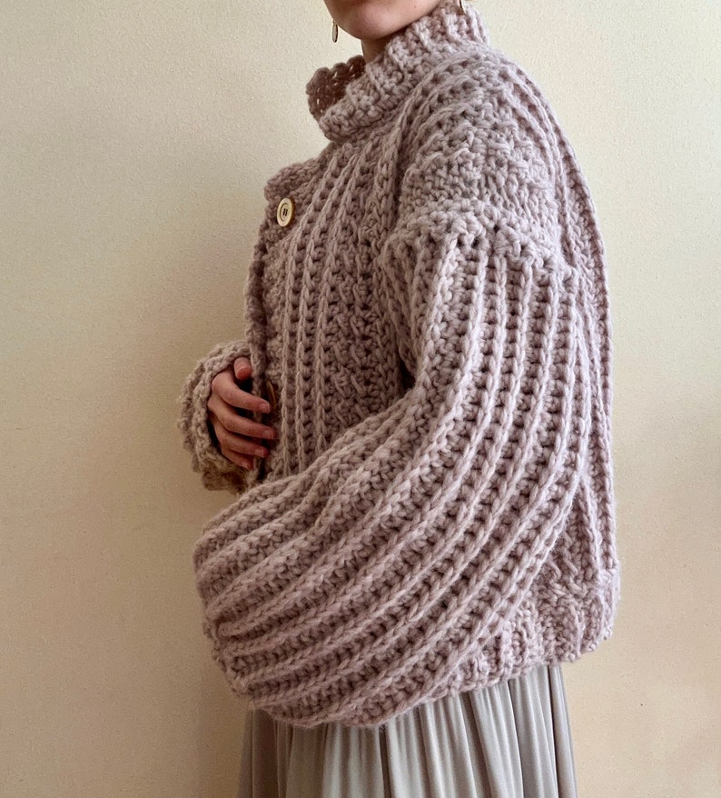 May include: A light pink crocheted cardigan with a high neck and long sleeves. The cardigan has a button closure and a textured stitch pattern.