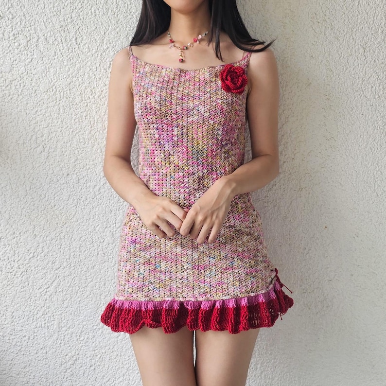 Puede incluir: Un vestido de crochet rosa y blanco con un dobladillo de volantes de crochet rojo y un adorno de flor roja.