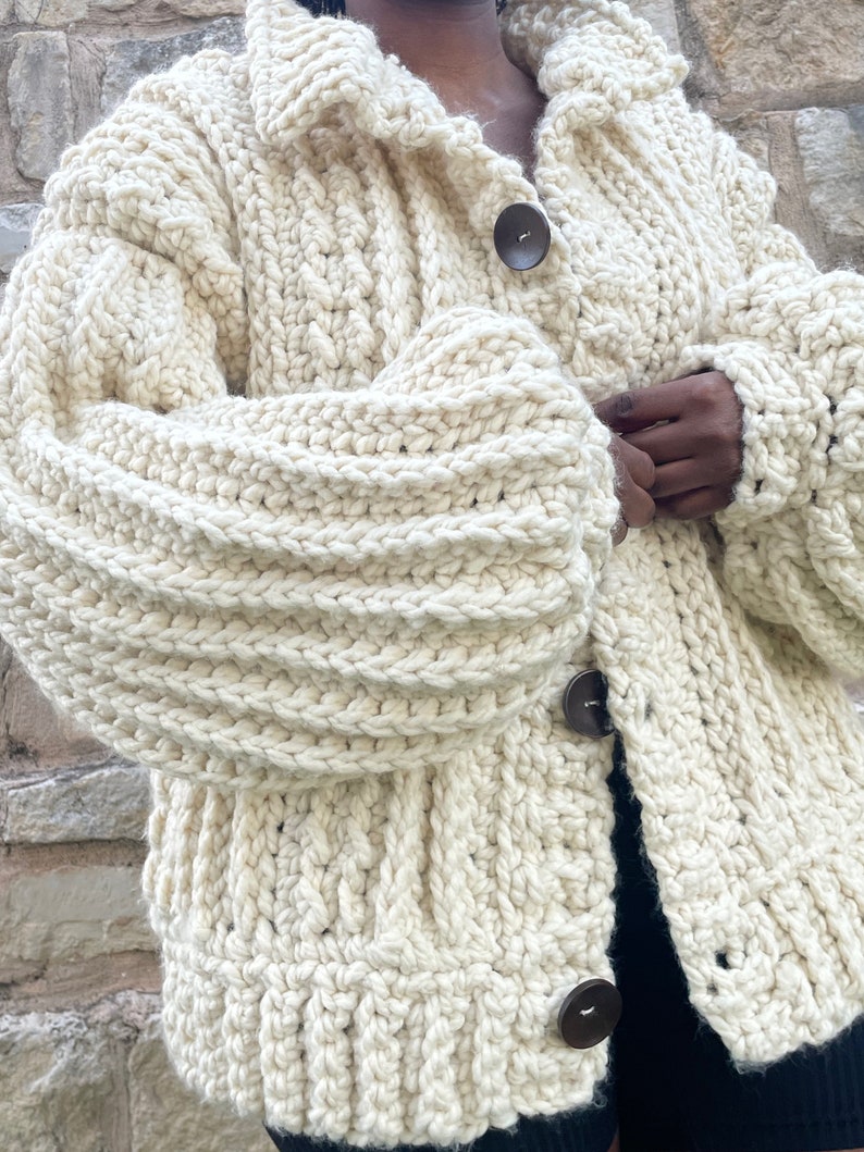 May include: A cream-colored crocheted cardigan with a collar and three brown buttons. The cardigan has a chunky knit texture.