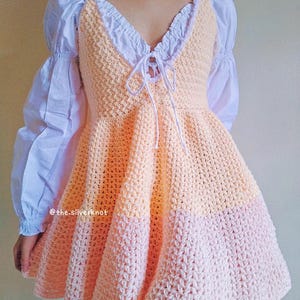 May include: A crochet dress with a peach and pink color gradient. The dress has a fitted bodice and a full skirt. The dress is tied at the neckline with a white ribbon.