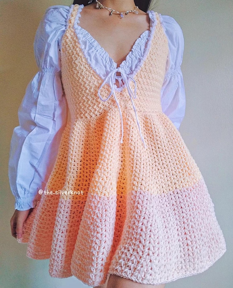 May include: A crochet dress with a peach and pink color gradient. The dress has a fitted bodice and a full skirt. The dress is tied at the neckline with a white ribbon.