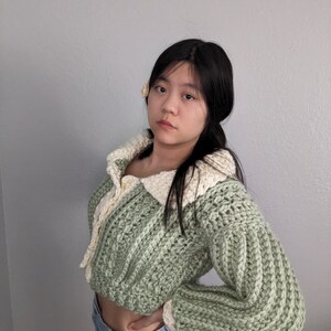 May include: A green and white crocheted sweater with a collared neckline and long sleeves. The sweater is cropped and has a ribbed texture.