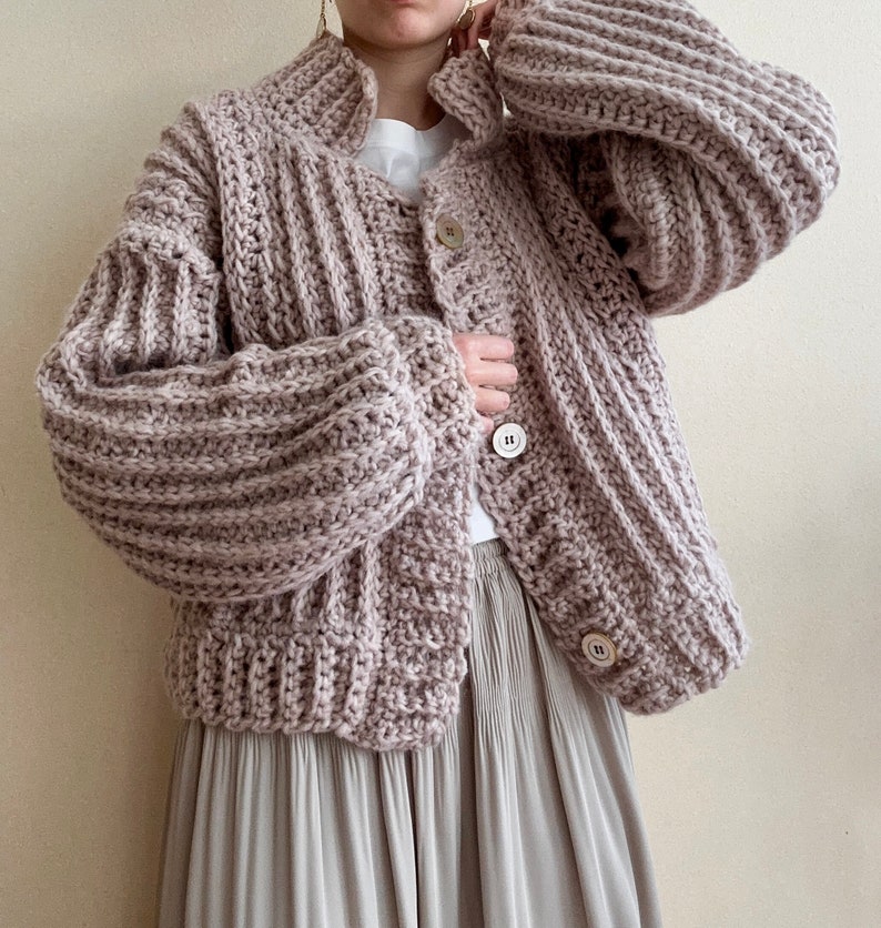 May include: A light pink knitted cardigan with a ribbed texture and button closure. The cardigan has long sleeves and a relaxed fit.