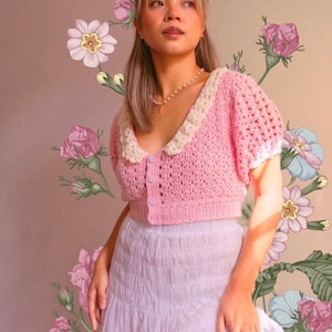 以下が含まれることがあります： 白い襟と短い袖が付いたピンクのクロシェのクロップトップ。トップは前面にボタン留めがあります。モデルは白いスカートと真珠のネックレスをしています。