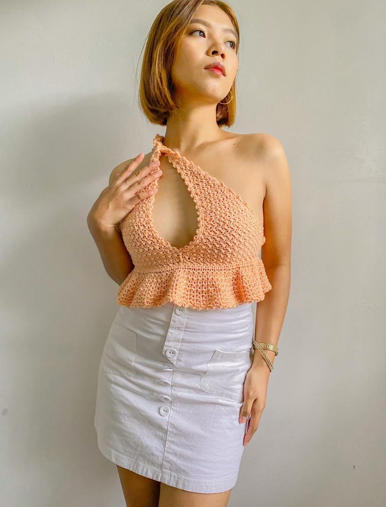 May include: A peach-colored crochet top with a one-shoulder design and a cutout detail. The top has a ruffled hem and is paired with a white mini skirt.