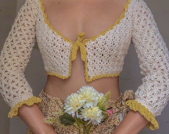 May include: A white crocheted crop top with yellow trim and a tie at the front. The top has a delicate floral pattern and is worn with a floral skirt.