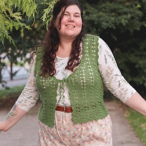 Puede incluir: Una mujer con un chaleco de crochet verde sobre una camisa de manga larga con estampado floral y pantalones florales. El chaleco tiene un cierre de lazo. Se lleva un cinturón marrón en la cintura. El fondo es una escena borrosa al aire libre.