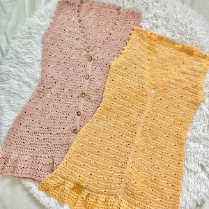 Op de afbeelding: Twee handgemaakte gehaakte jurken, één zachtroze en de andere zonnegeel. Beide hebben een V-hals, knoopsluiting en een gefranjerde zoom. De jurken zijn mouwloos en lijken van lichtgewicht garen te zijn.