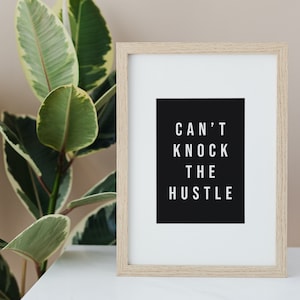 Peut inclure: Une impression encadrée avec un fond noir et du texte blanc qui dit "CAN'T KNOCK THE HUSTLE". Le cadre est en bois clair et l'impression est posée sur une surface blanche.