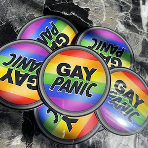 Może przedstawiać: Pięć okrągłych naklejek z tęczowym wzorem i napisem "Gay Panic" czarnymi literami. Naklejki są ułożone jedna na drugiej.