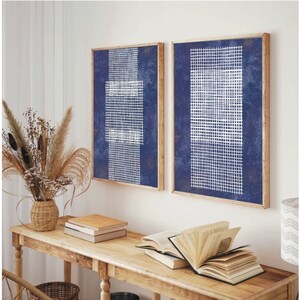 May include: Two framed abstract art prints with a blue background and white grid patterns. The prints are hanging on a white wall above a wooden console table with books and a vase of dried flowers.
