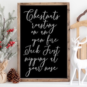 May include: Black framed sign with white text that reads "Chestnuts roasting on an open fire Jack Frost nipping at your nose". The sign is sitting on a white surface with a small snowy pine tree and a white deer figurine.